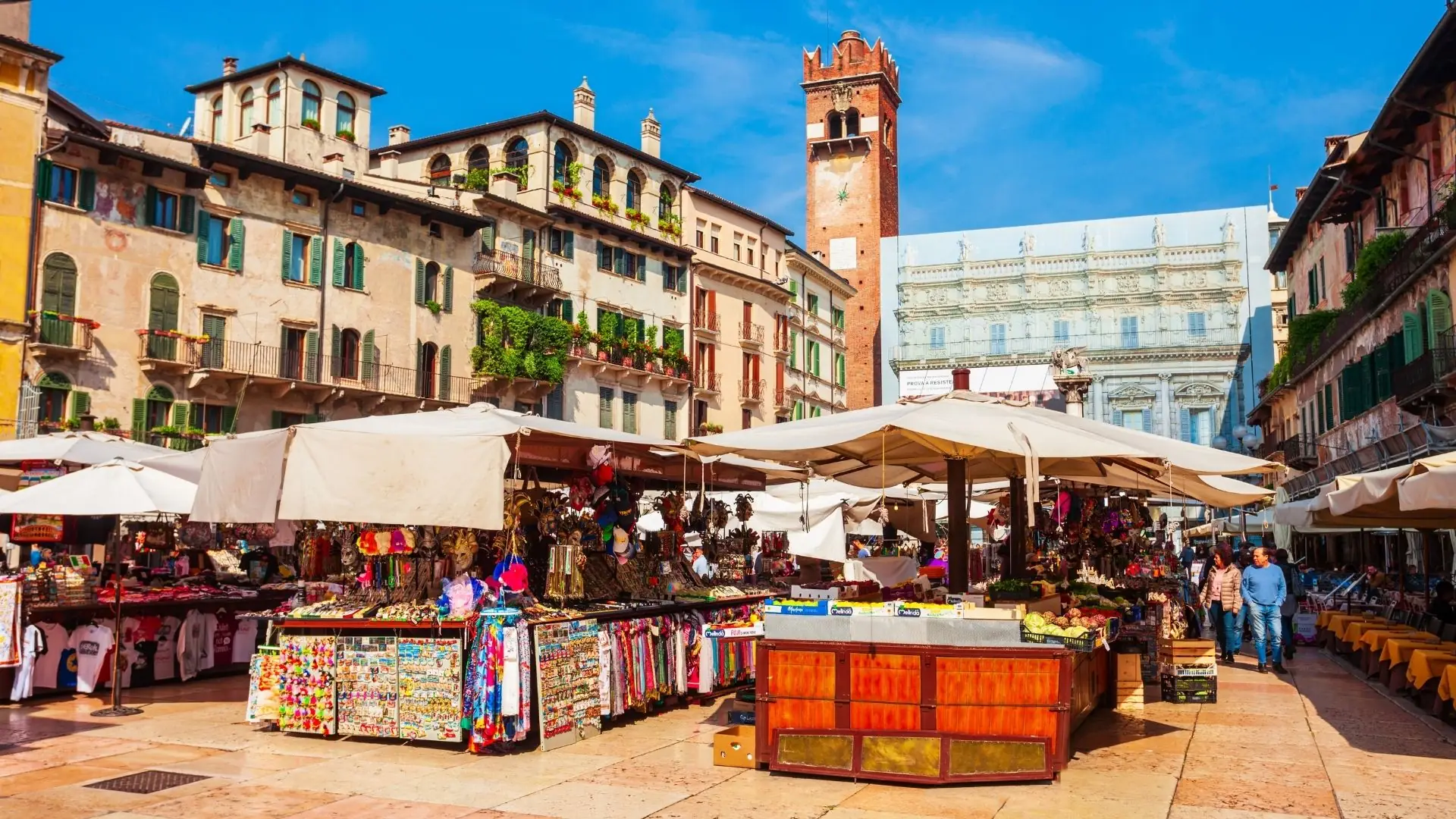 Marktstände mit bunten Waren und weißen Sonnenschirmen auf dem belebten Platz Piazza delle Erbe in Verona, umgeben von historischen Gebäuden und einem hohen Turm.