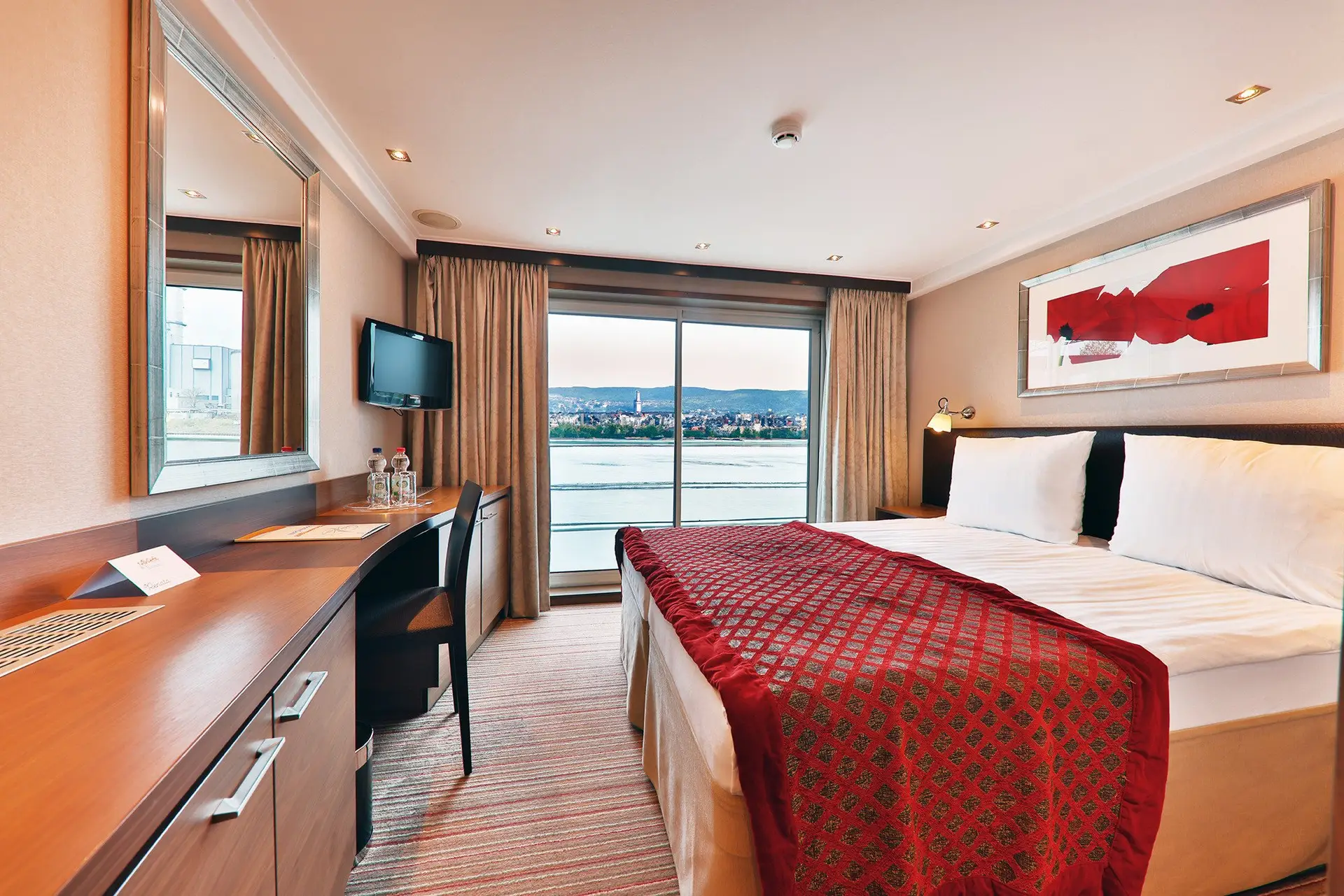 Hotelzimmer mit Doppelbett, roter Tagesdecke, Schreibtisch, Fernseher an der Wand und großem Fenster mit Blick auf einen Fluss und Stadt im Hintergrund