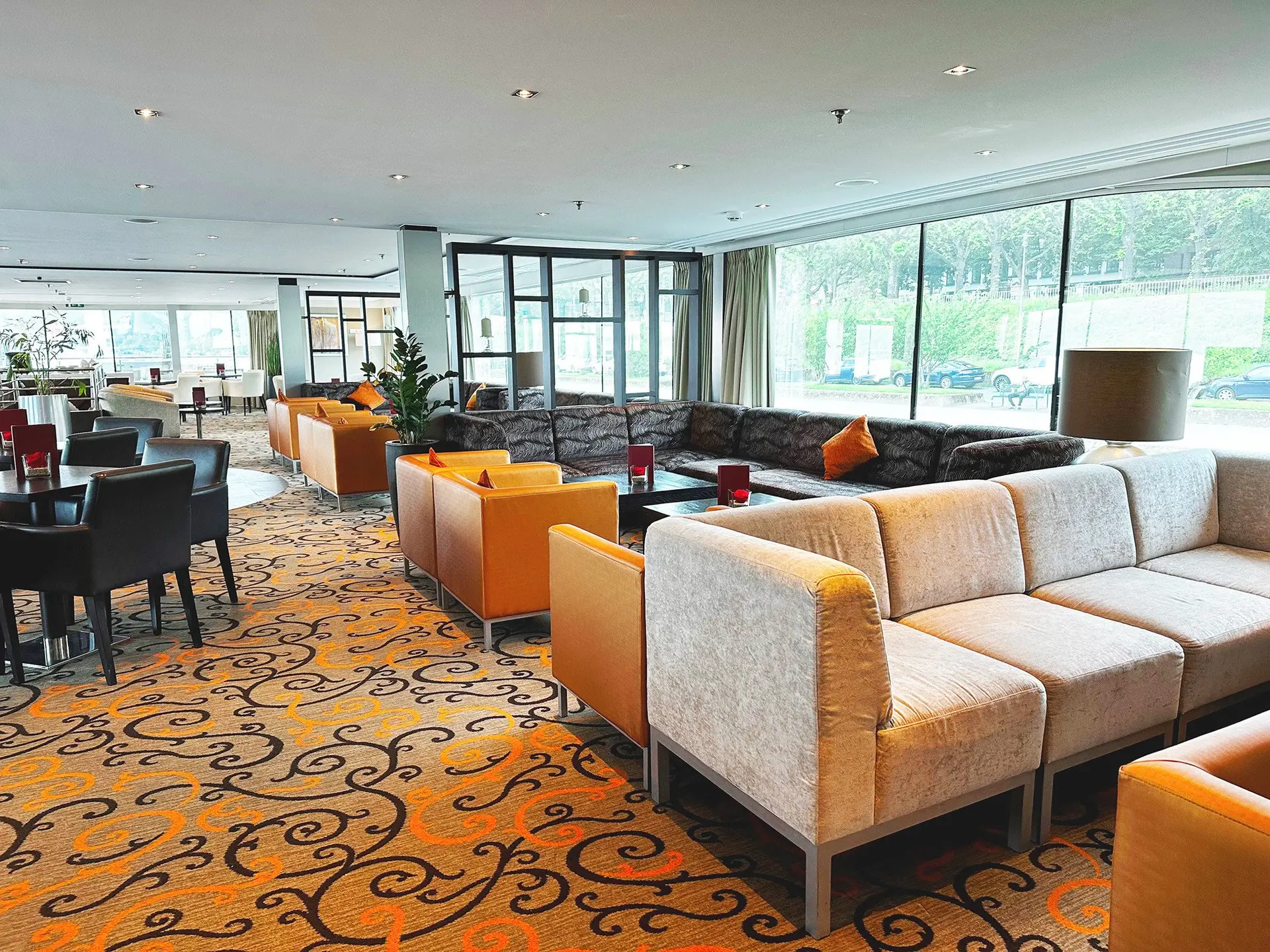 Großer, heller Aufenthaltsraum mit Sofas in Beige und Orange, Teppich mit geschwungenem Muster und großen Fenstern zur Gartenseite