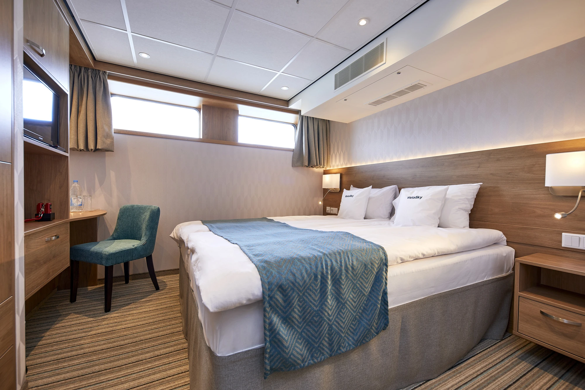 Doppelbett mit weißen Kissen und blauem Läufer, grüner Stuhl, Holzschreibtisch, Fenster mit Vorhängen, Wandlampen und Teppichboden in einer Kabine.