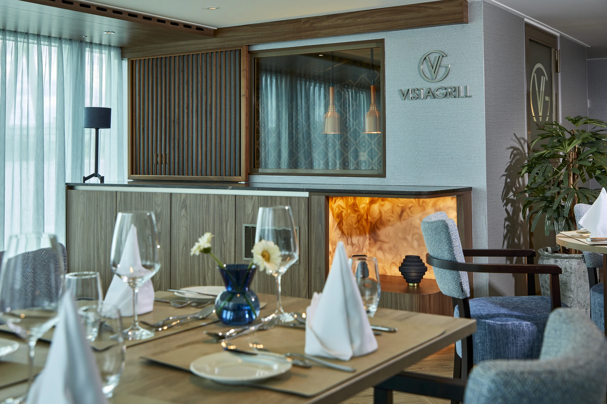 Elegantes Restaurantinterieur mit gedecktem Holztisch, weißen Servietten, Gläsern, blauer Vase mit Blume und Sitzgelegenheiten aus Stoff.