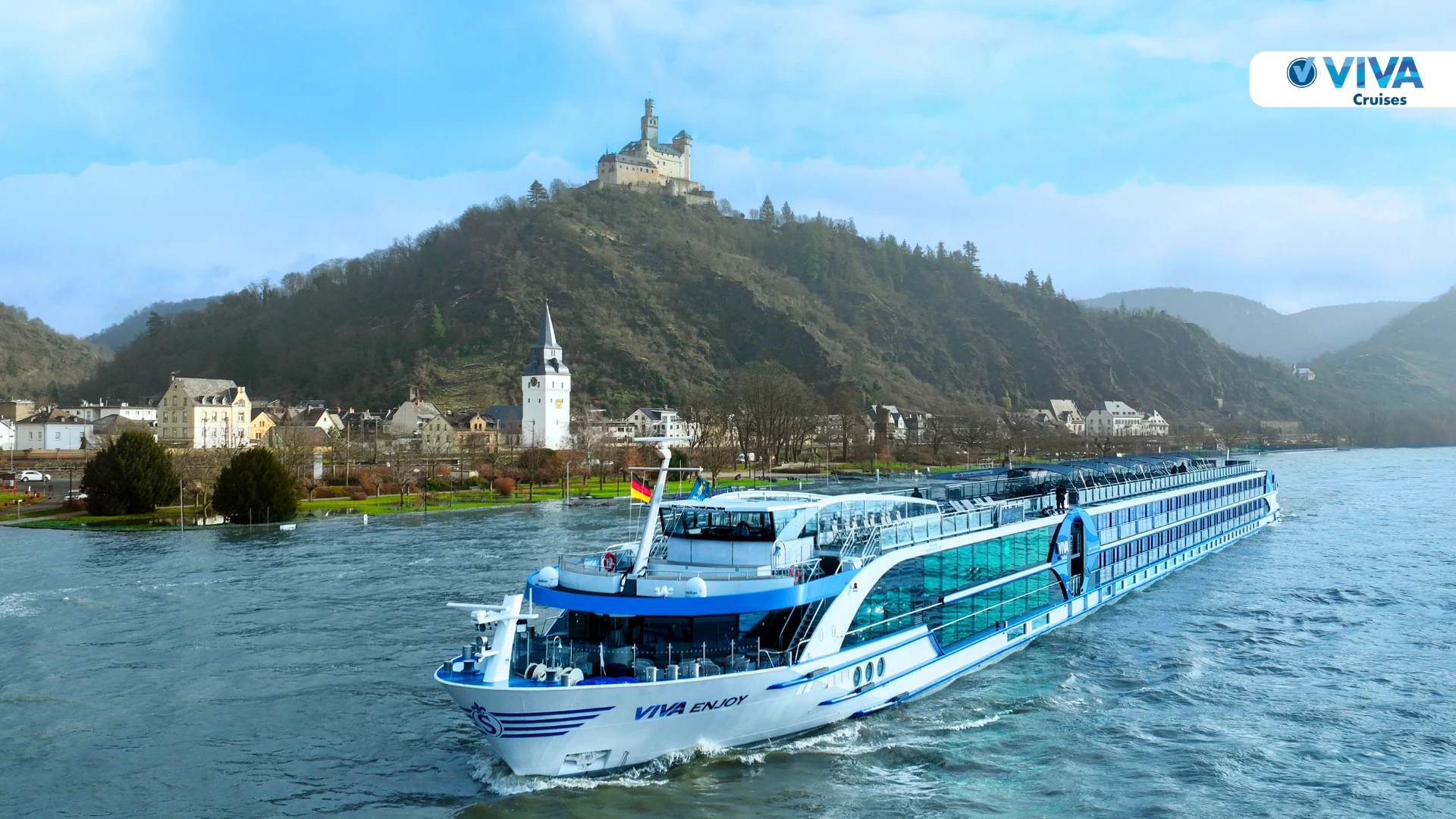 Flusskreuzfahrtschiff VIVA Enjoy fährt auf einem Fluss mit einer Stadt und einem bewaldeten Hügel mit einer Burg im Hintergrund