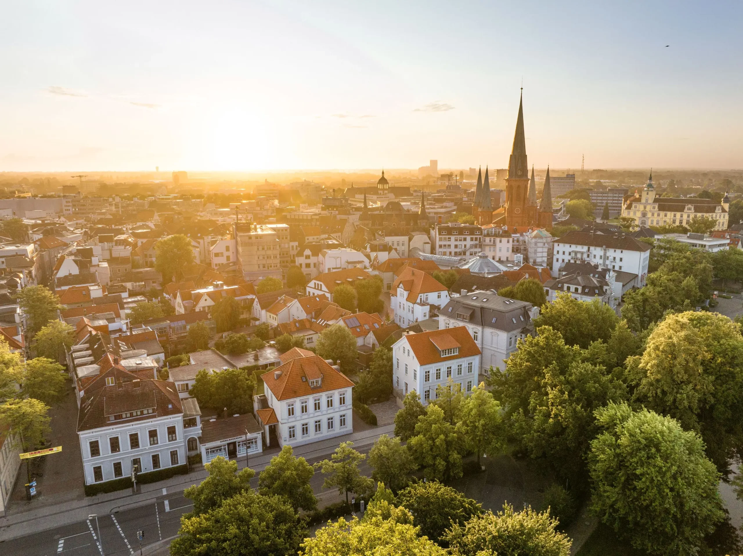 Vogelperspektive auf die Stadt Oldenburg mit vielen Gebäuden, einer großen Kirche mit hohem Turm und viel Grün bei Sonnenuntergang