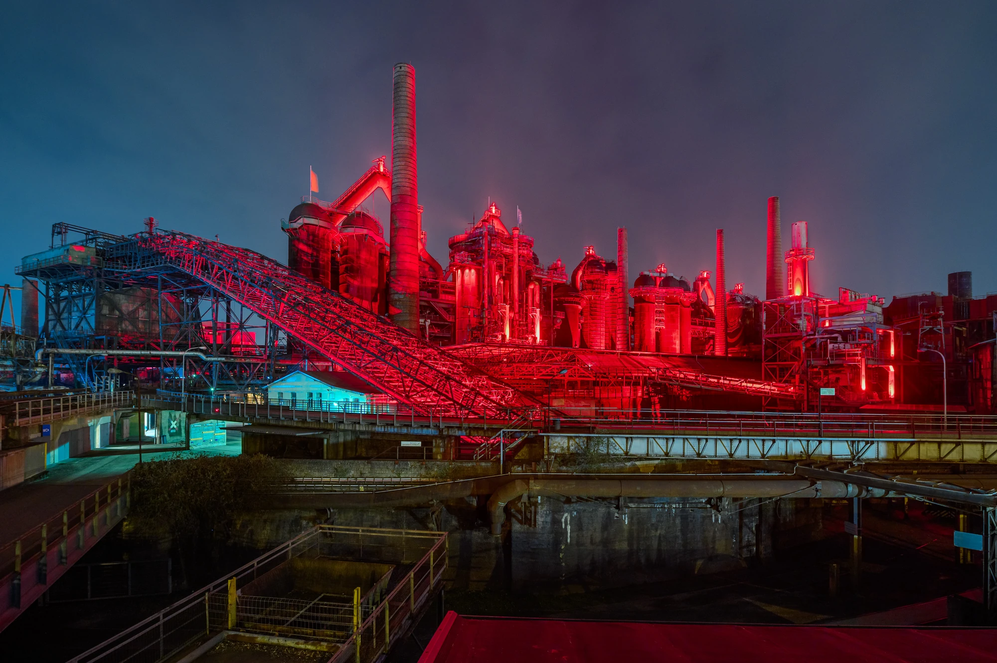 Industrieanlage Völklinger Hütte bei Nacht, beleuchtet in rotem Licht, mit Hochöfen, Rohrleitungen und Förderbändern.