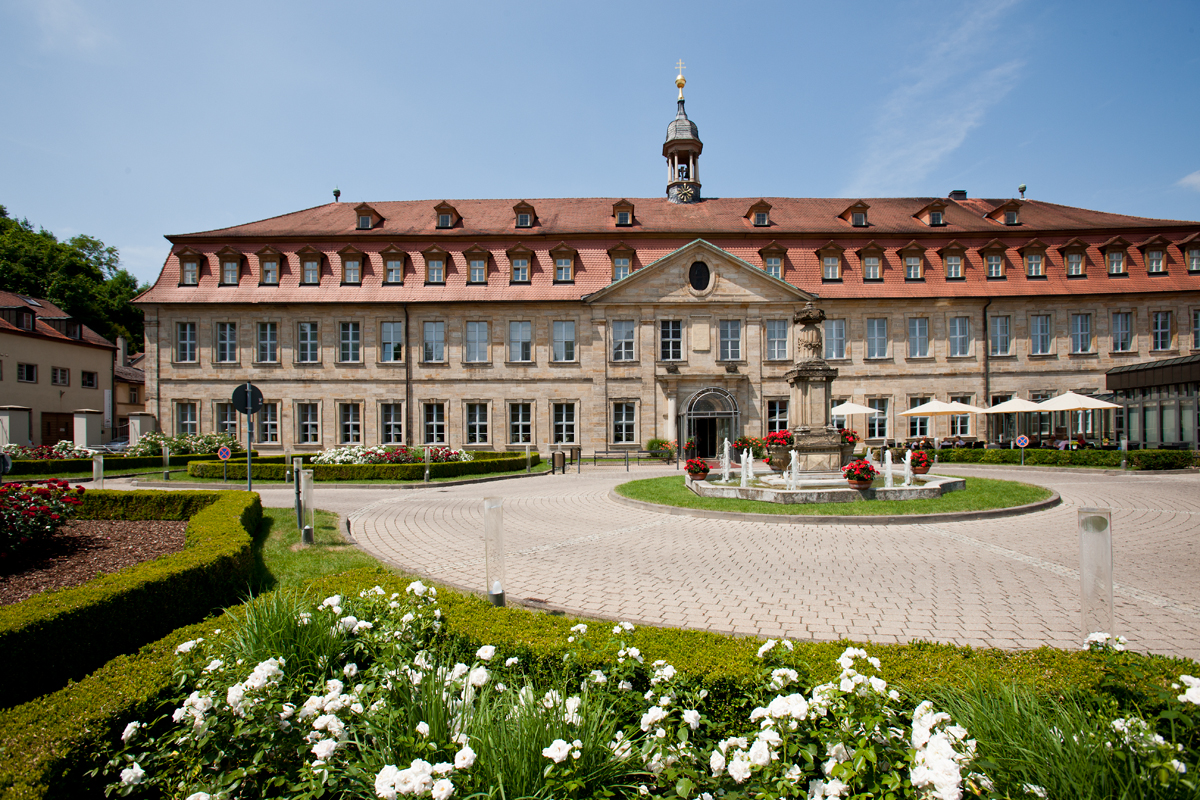 Breitformatige Außenansicht des historischen Residenzschlosses in Bamberg mit rotem Ziegeldach, symmetrischer Fassade und einem zentralen Eingangsportal, davor ein Brunnen und bepflanzte Beete