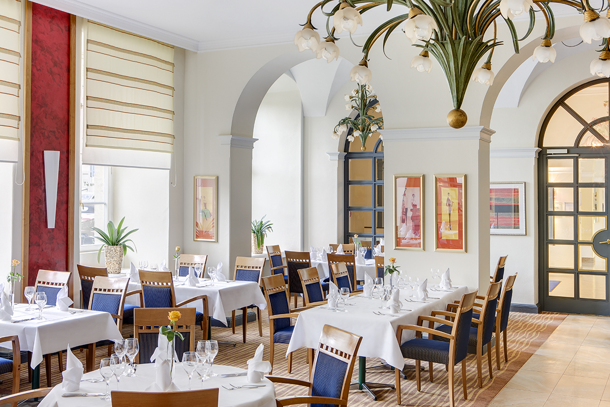 Elegantes Restaurant mit weißen Tischdecken, blauen Stühlen, großen Fenstern, dekorativen Pflanzen und Kunstwerken an den Wänden im Residenzschloss Bamberg