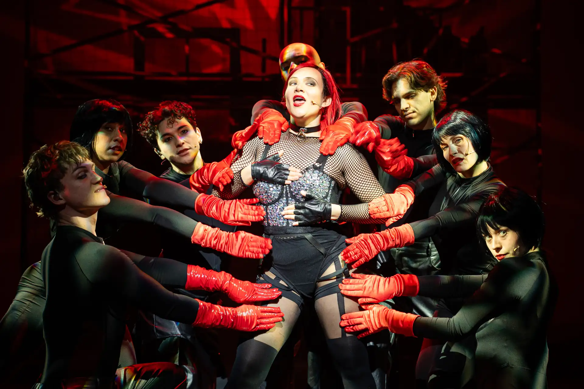 Eine Frau in Netzstrumpfhose und schwarzem Outfit wird von mehreren Personen mit schwarzen Oberteilen und roten Handschuhen umgeben, die ihre Arme und Hände auf sie legen.