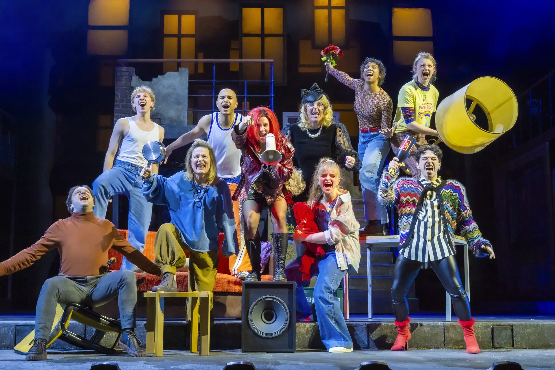 Theaterensemble mit neun Personen in bunter Kleidung auf einer Bühne mit Treppen und Fenstern im Hintergrund, einige halten Requisiten wie einen Lautsprecher und eine Blume