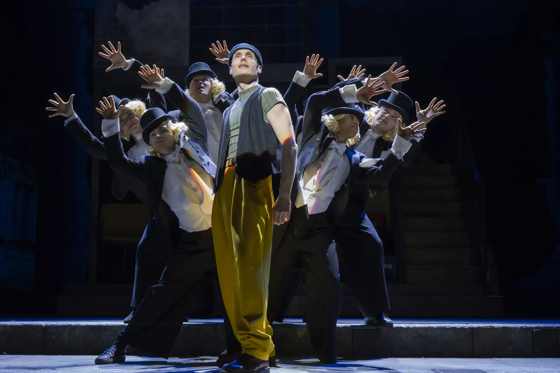 Gruppe von sieben Tänzern auf Bühne mit erhobenen Händen, vorn ein Tänzer in gelben Hosen und Weste