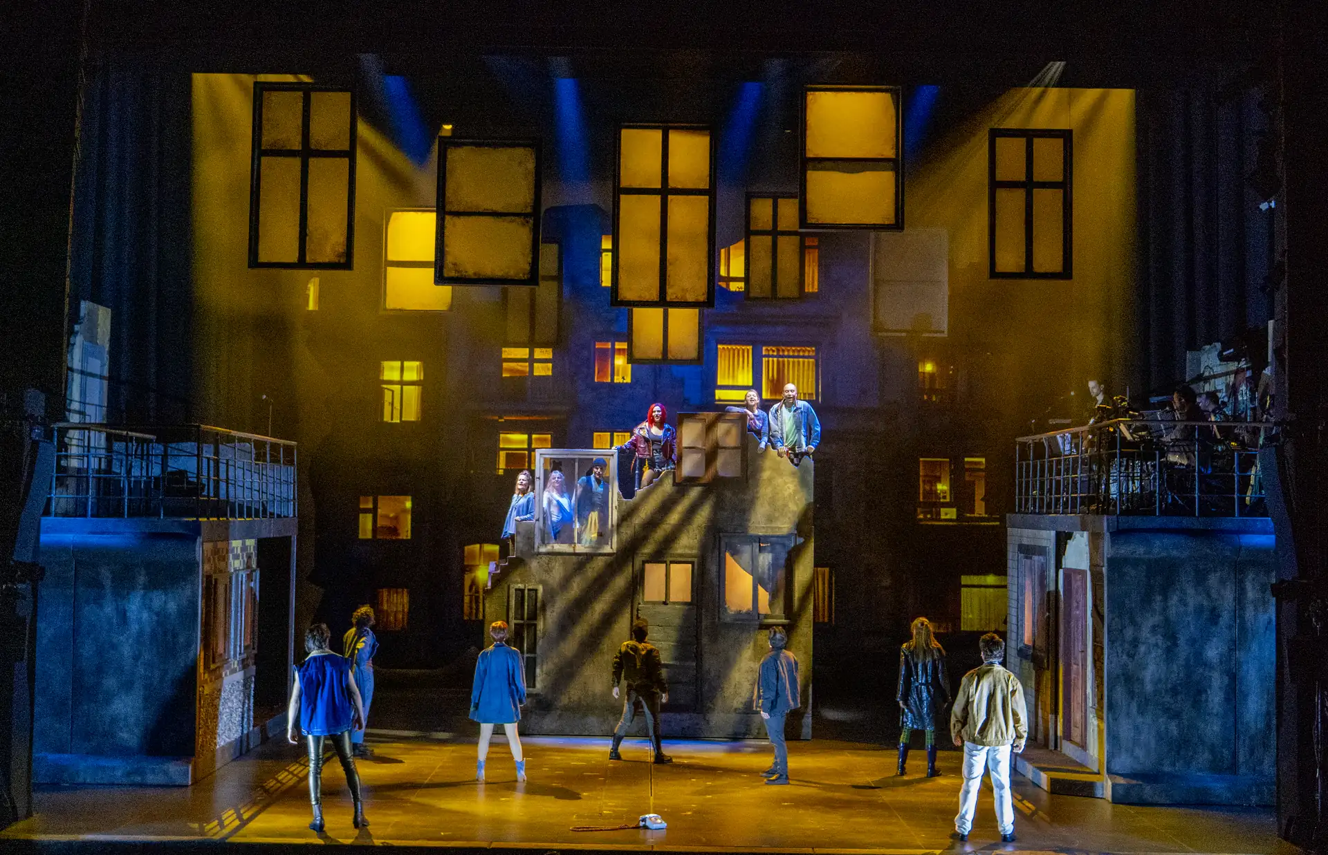 Bühnenbild mit mehreren Schauspielern vor einer Kulisse aus beleuchteten Fenstern und hängenden Fensterrahmen in einem Theaterstück