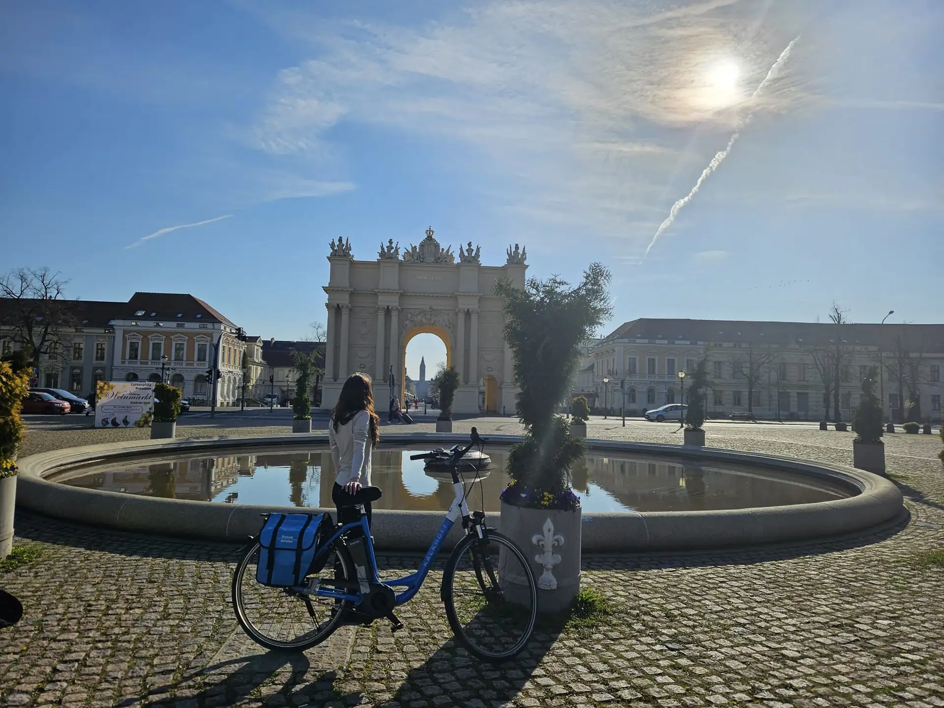 Frau mit Fahrrad steht vor einem runden Brunnen, im Hintergrund das Kleine Brandenburger Tor in Potsdam bei sonnigem Himmel