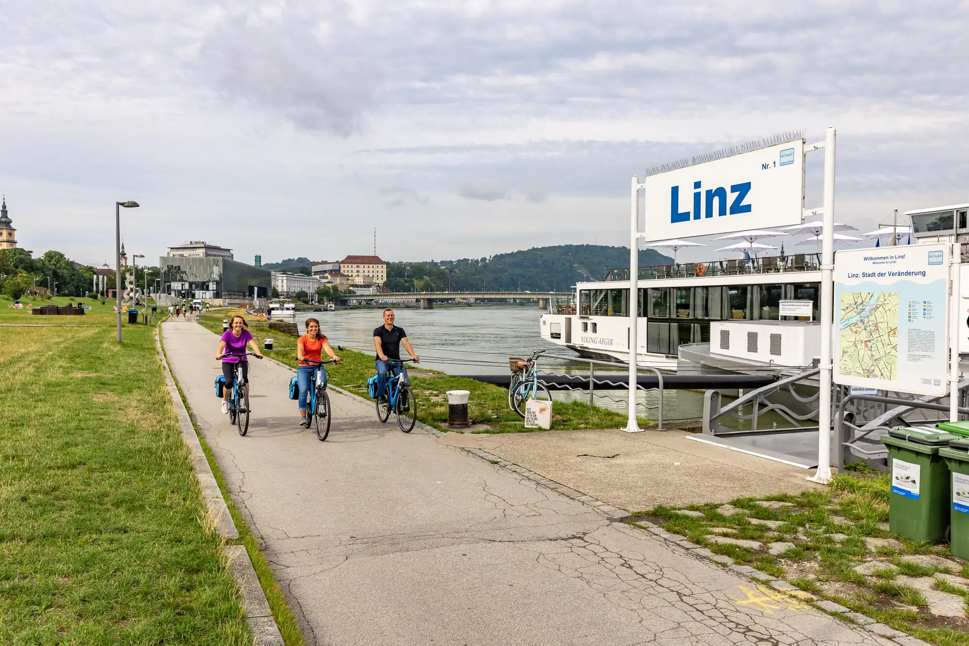 Drei Radfahrer fahren auf dem Donauradweg an Anlegestelle in Linz vorbei.