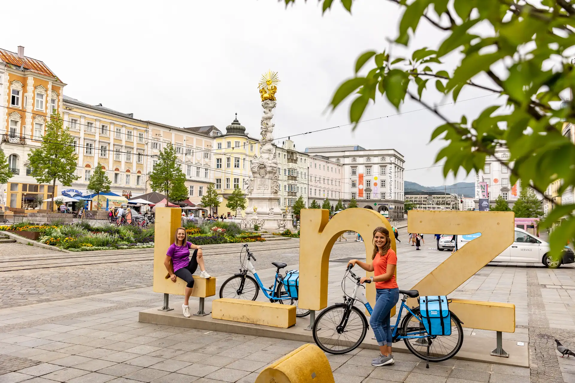 Zwei fröhliche Frauen mit Rädern am Schriftzug Linz am Hauptplatz.