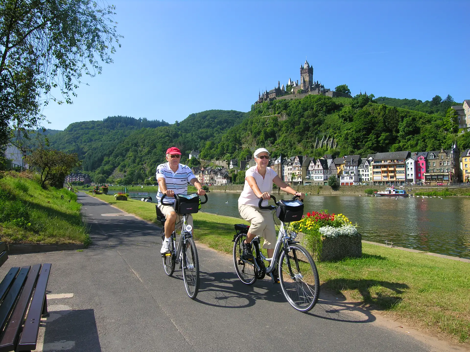 Älteres Paar auf Rädern am Ufer der Mosel mit im Hintergrund thronender Reichsburg bei Sonnenschein