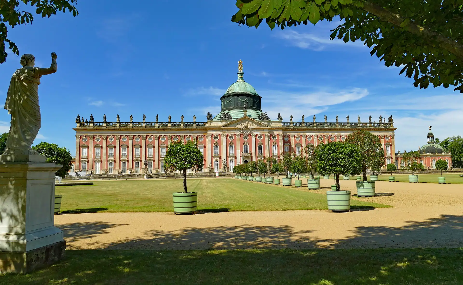 Neues Palais in Potsdam mit rotem Fassadenfront, grüner Kuppel und Statuen auf dem Dach, davor Garten mit Bäumen in Pflanzkübeln und Statue links