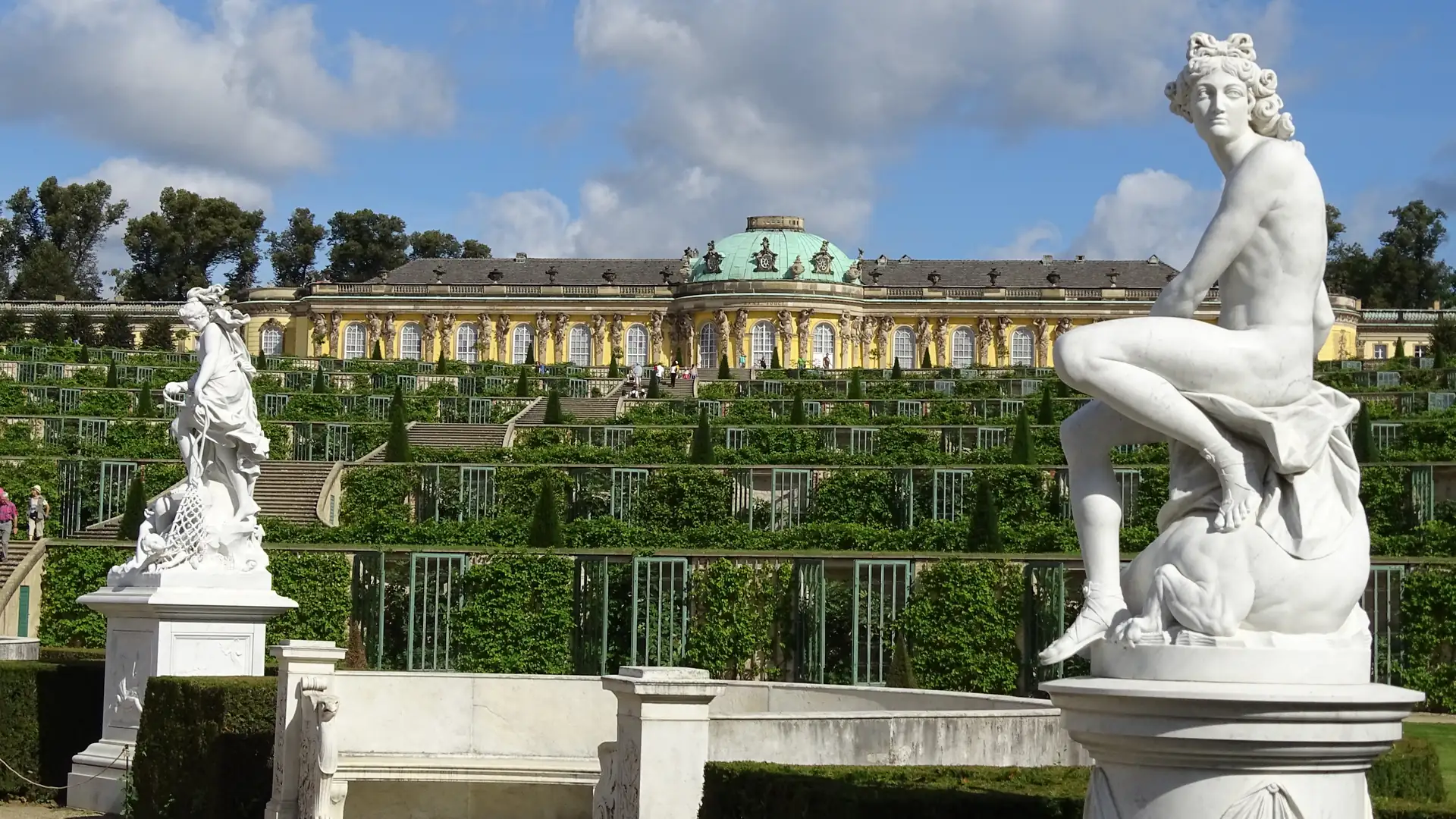 Schloss Sanssouci in Potsdam mit terrassiertem Garten und zwei weißen Statuen im Vordergrund unter blauem Himmel