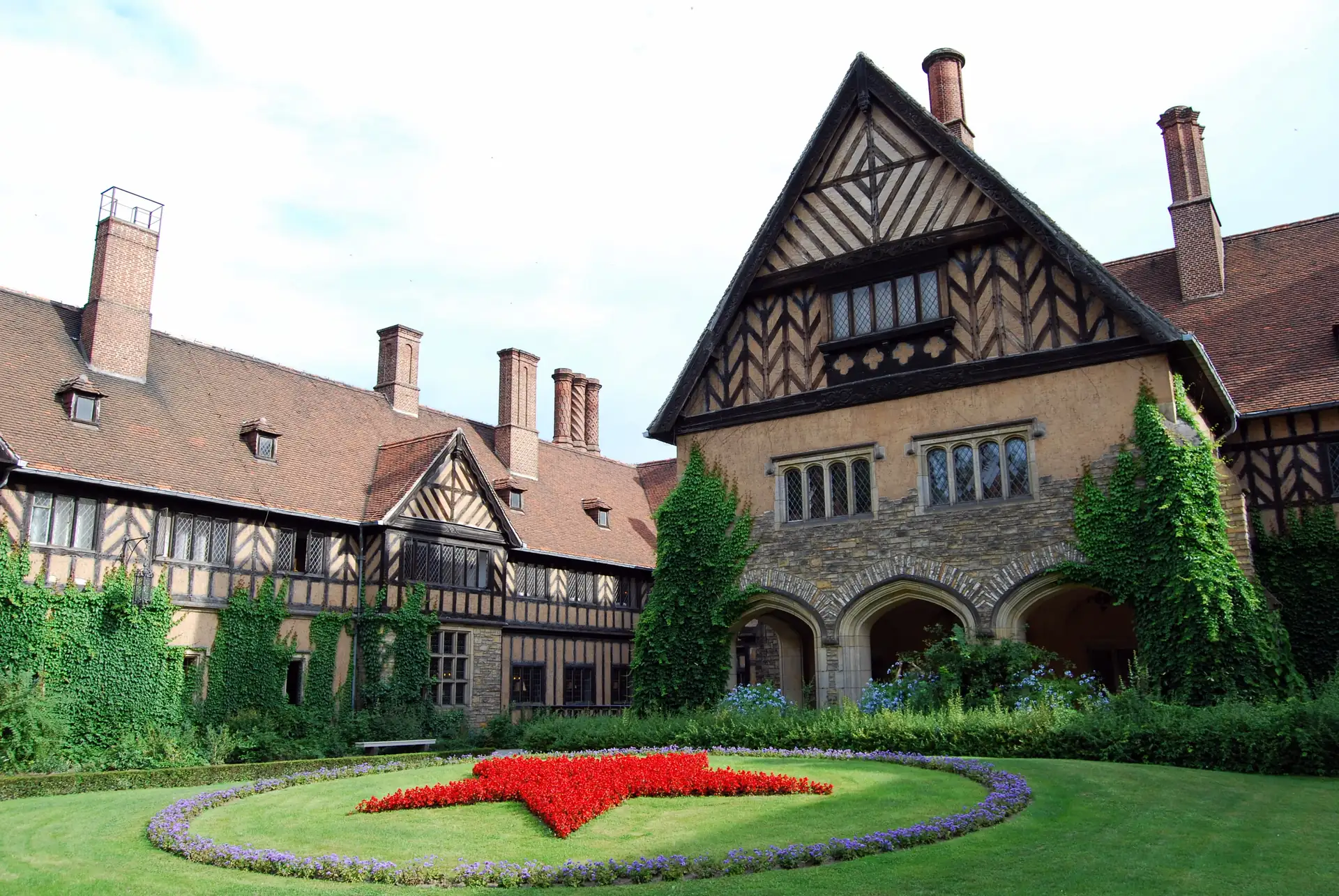 Historisches Fachwerkgebäude mit roten Ziegeldächern und mehreren Kaminen, davor ein Garten mit einem großen roten Stern aus Blumen umgeben von lila Blumen