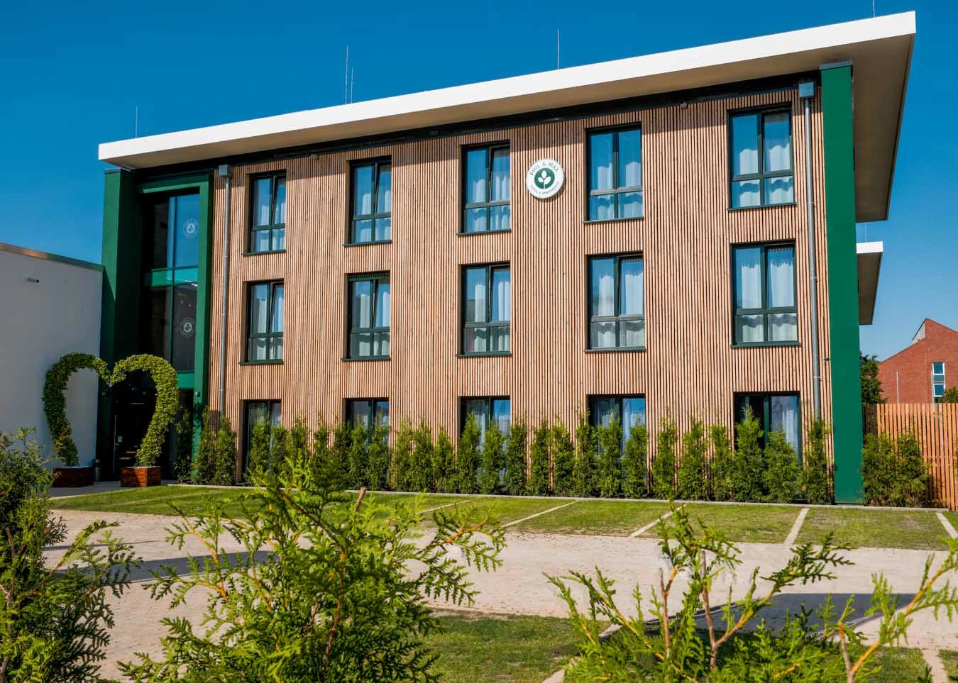 Das Bild zeigt die Fassade des Phil & Max Hotels in modernem, zeitgemäßem Design. Die stilvolle Holzverkleidung harmoniert mit den grünen Akzenten, die dem Gebäude einen natürlichen und einladenden Charakter verleihen. Die großen Fenster ermöglichen viel Tageslicht und sorgen für eine offene Atmosphäre. Der Eingangsbereich wird durch einen dekorativen, herzförmigen Bogen aus Pflanzen hervorgehoben, was dem Hotel einen charmanten und besonderen Touch verleiht. Vor dem Gebäude erstreckt sich ein gepflegter Garten mit dezent angelegten Wegen, umrahmt von Hecken, die Privatsphäre und ein ruhiges Ambiente schaffen. Das Phil & Max Hotel präsentiert sich hier als modernes, einladendes Domizil mit hohem ästhetischem Anspruch.