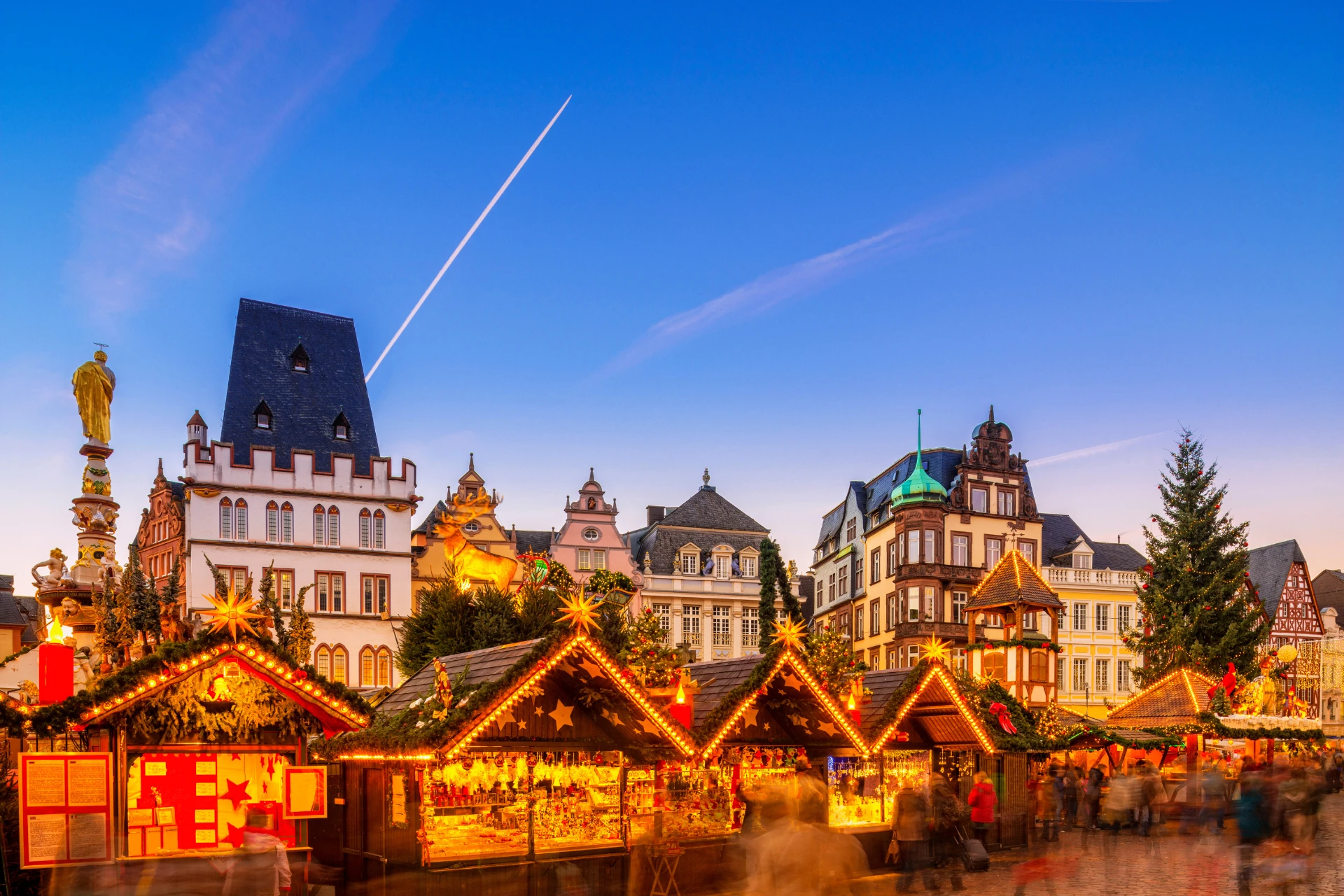 Weihnachtsmarkt mit beleuchteten Holzhütten vor historischen Gebäuden und einem großen Weihnachtsbaum in Trier bei Abendlicht