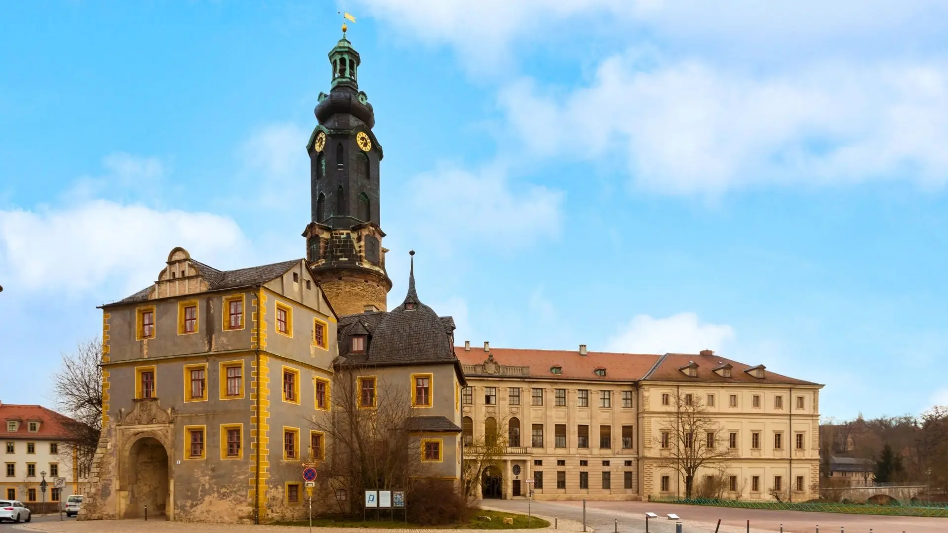 Das Weimar Stadtschloss zeigt eine historische Architektur mit einem hohen, runden Turm und einem rechteckigen Gebäude im Vordergrund. Der Turm hat eine goldene Fahne und ist mit einem Ziffernblatt versehen. Im Hintergrund sind weitere Gebäude mit roten Dächern zu sehen.