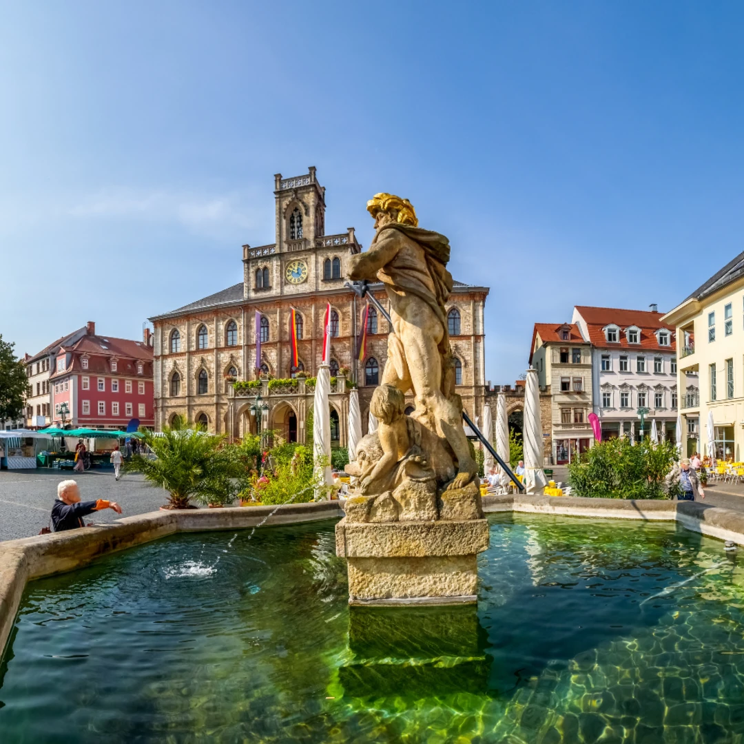 Dieses Foto zeigt den Marktplatz in Weimar mit dem Neptunbrunnen im Vordergrund. Im Hintergrund steht das historische Rathaus mit seiner markanten Fassade und der Turmuhr. Das lebendige Stadtbild mit Marktständen und Cafés verleiht der Szene eine einladende Atmosphäre.