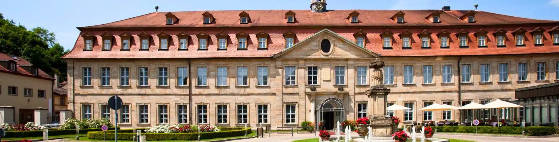 Breitformatige Außenansicht des historischen Residenzschlosses in Bamberg mit rotem Ziegeldach, symmetrischer Fassade und einem zentralen Eingangsportal, davor ein Brunnen und bepflanzte Beete