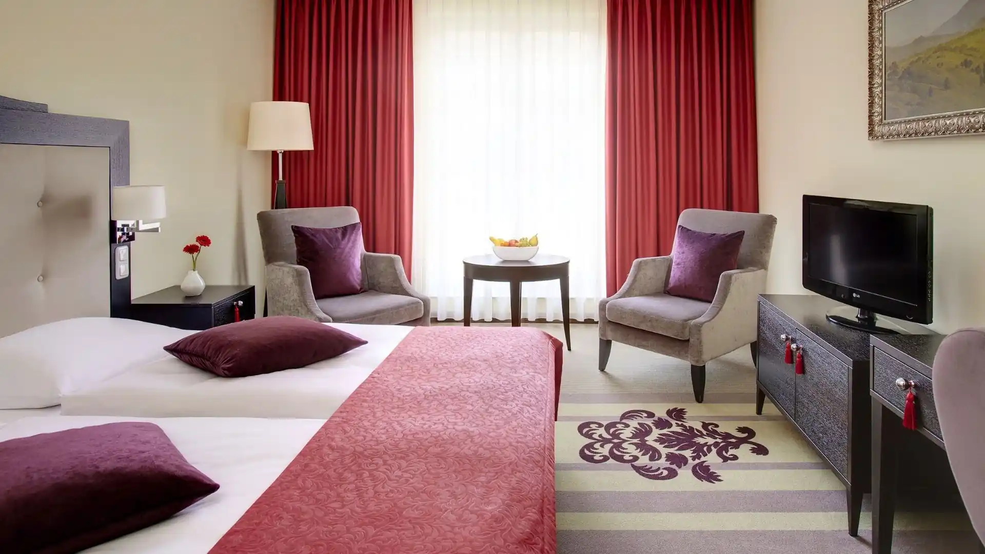 Hotelzimmer mit zwei Einzelbetten, roten Bettläufern und Kissen, zwei grauen Sesseln mit lila Kissen, kleinem runden Tisch, Fernseher auf Sideboard und roten Vorhängen vor hellem Fenster