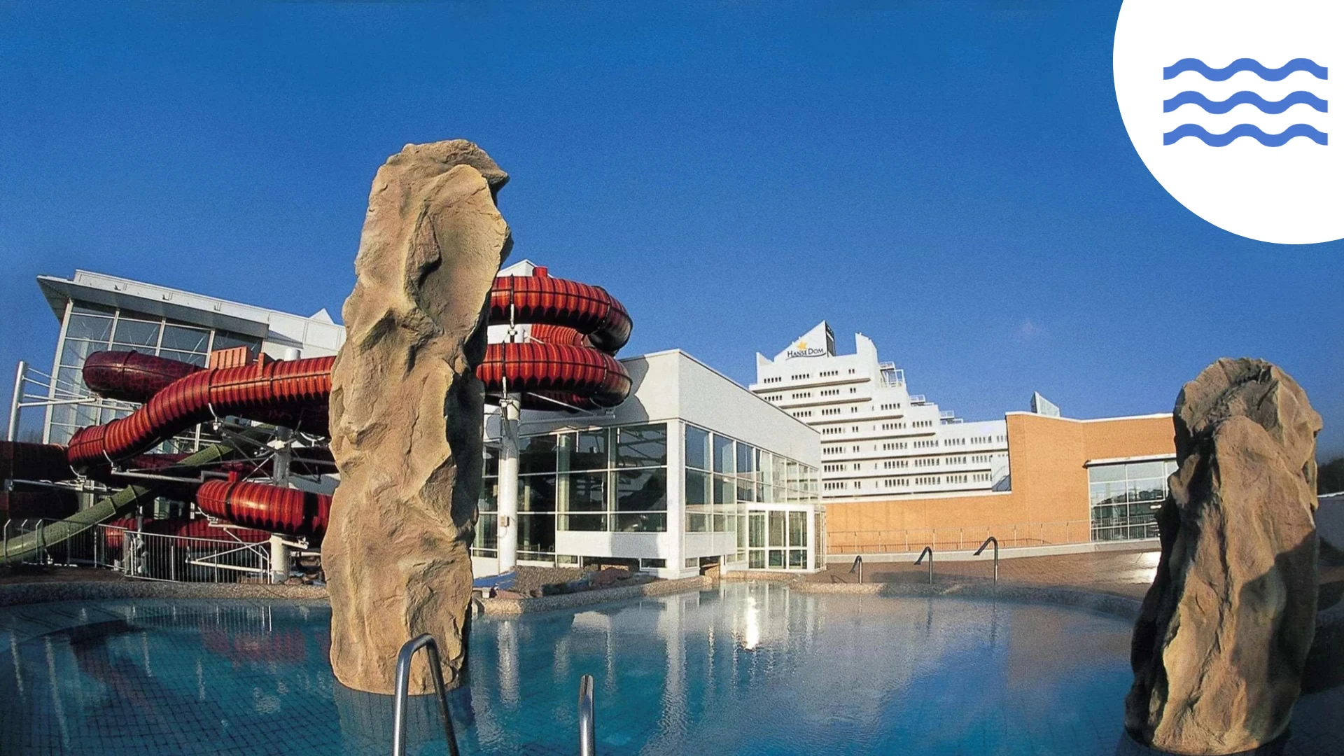 Ein lebendiger Wasserpark mit einer Felsenskulptur, einem Schwimmbecken und roten Wasserrutschen unter einem strahlend blauen Himmel.