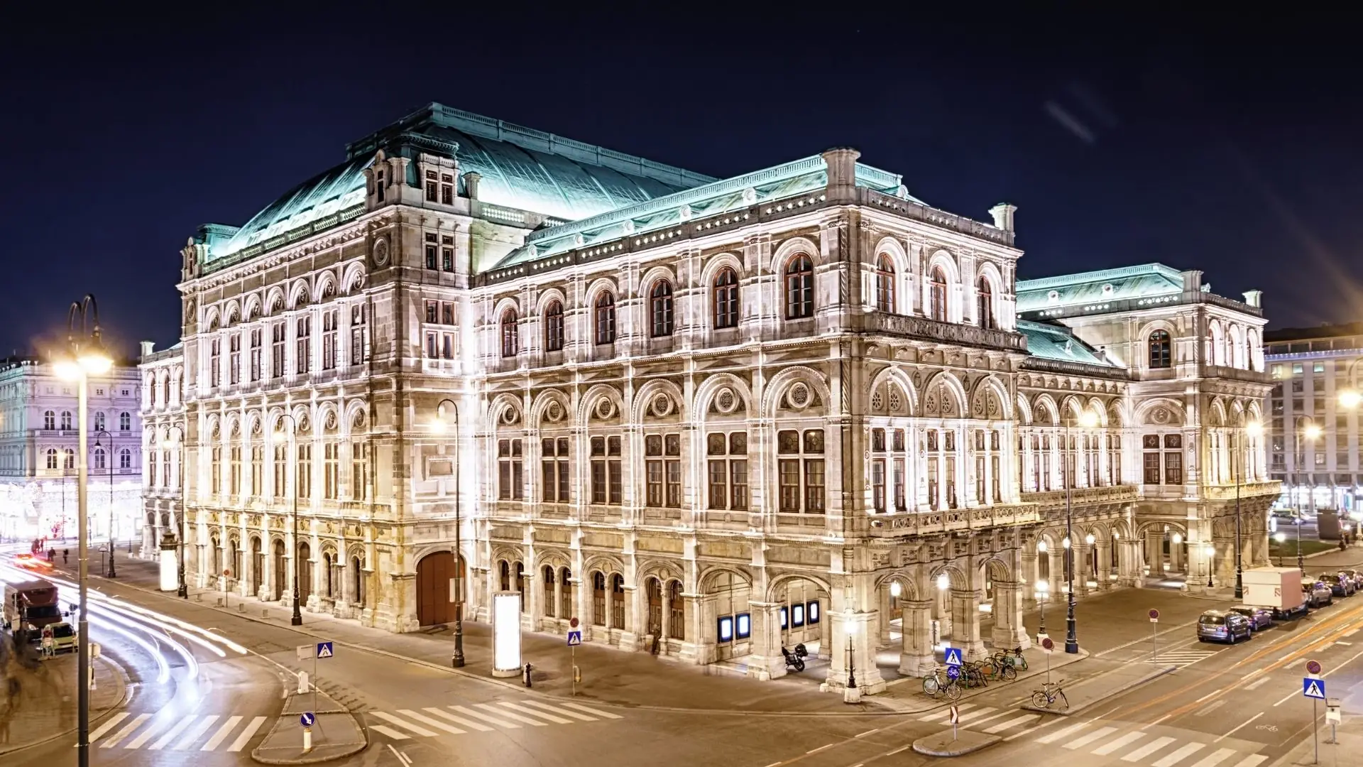 Beleuchtetes historisches Gebäude der Wiener Staatsoper bei Nacht an einer Straßenkreuzung mit Verkehrsschildern und geparkten Autos.
