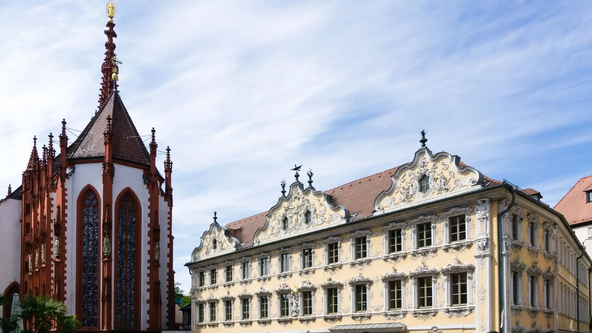 Barockes Gebäude mit reich verzierten Fassaden und spitzem Turm in Würzburg bei bewölktem Himmel.
