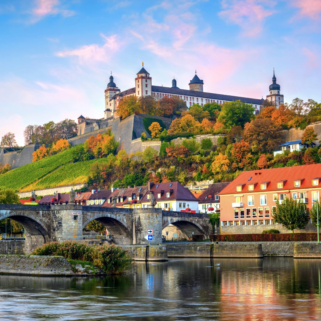 Dieses Foto zeigt die Festung Marienberg, die majestätisch über Würzburg thront. Im Vordergrund fließt der Main mit der Alten Mainbrücke, die von historischen Gebäuden umgeben ist. Die herbstlichen Farben der Weinberge verleihen der Szene eine besondere Atmosphäre.