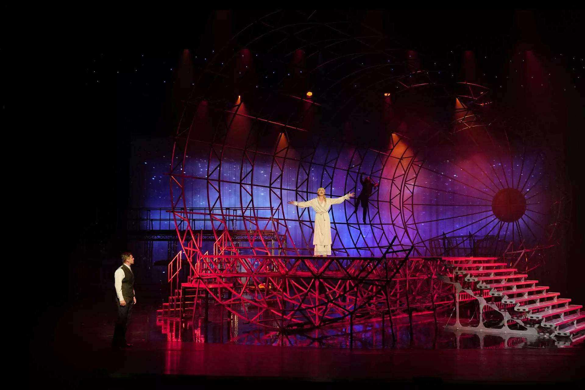 Musical Zeppelin: Szene mit zwei Schauspielern auf einer roten, runden Metallkonstruktion, einer steht oben mit ausgebreiteten Armen in weißem Kostüm, der andere unten in dunkler Kleidung