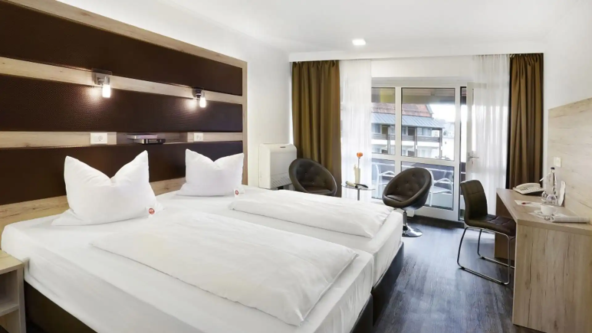 Hotelzimmer mit zwei Einzelbetten, zwei Sesseln vor bodentiefem Fenster mit Balkon, Schreibtisch mit Stuhl und moderner Beleuchtung