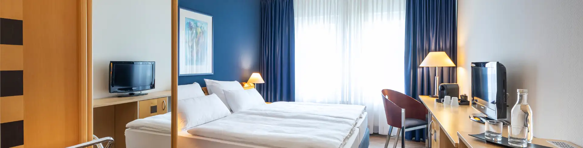 Achat Dresden Elbufer: Hotelzimmer mit zwei Betten, Schreibtisch mit Stuhl, Fernseher und Lampe vor einem Fenster mit blauen Vorhängen.