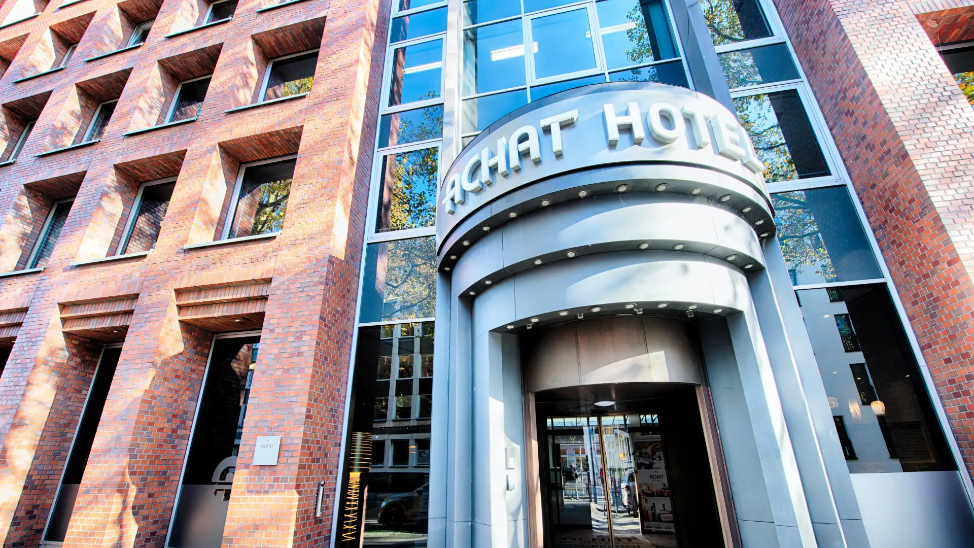 Eingang des ACHAT Hotels in Bremen mit einem runden, silbernen Vordach und großen Glasfenstern. Die Fassade ist aus roten Ziegelsteinen.