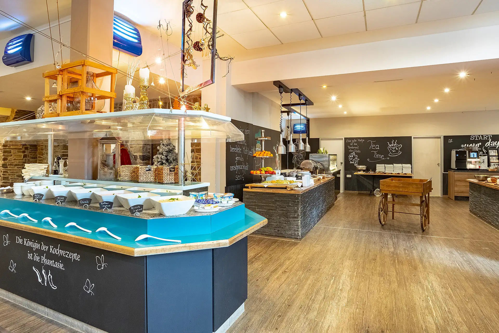 Buffetbereich mit verschiedenen Frühstücksangeboten in einem hellen Hotelraum mit Holzboden und dekorativen Tafeln an den Wänden