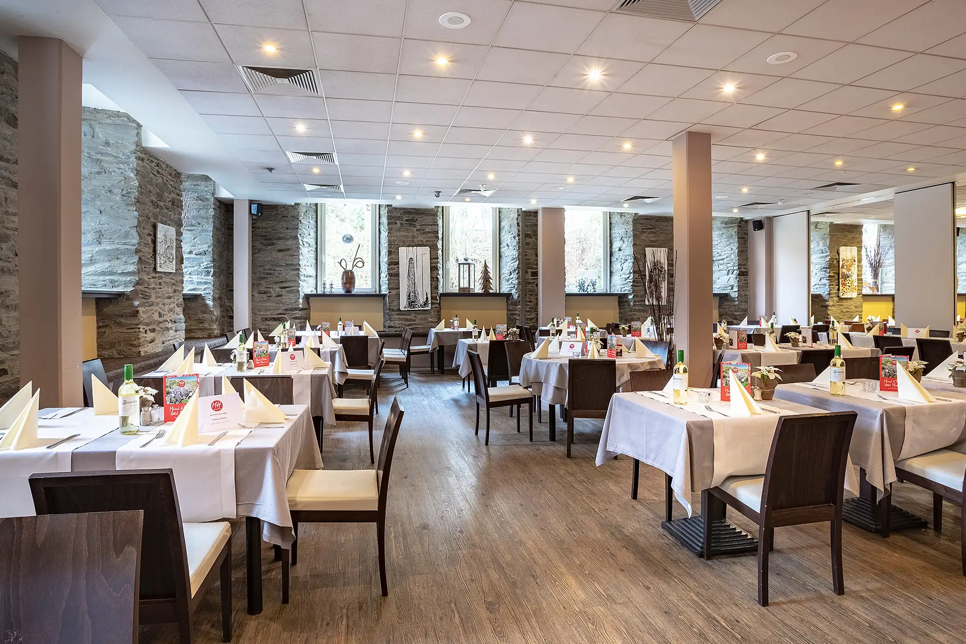 Restaurant mit mehreren eingedeckten Tischen und Stühlen in einem hellen Raum mit Steinwänden und großen Fenstern