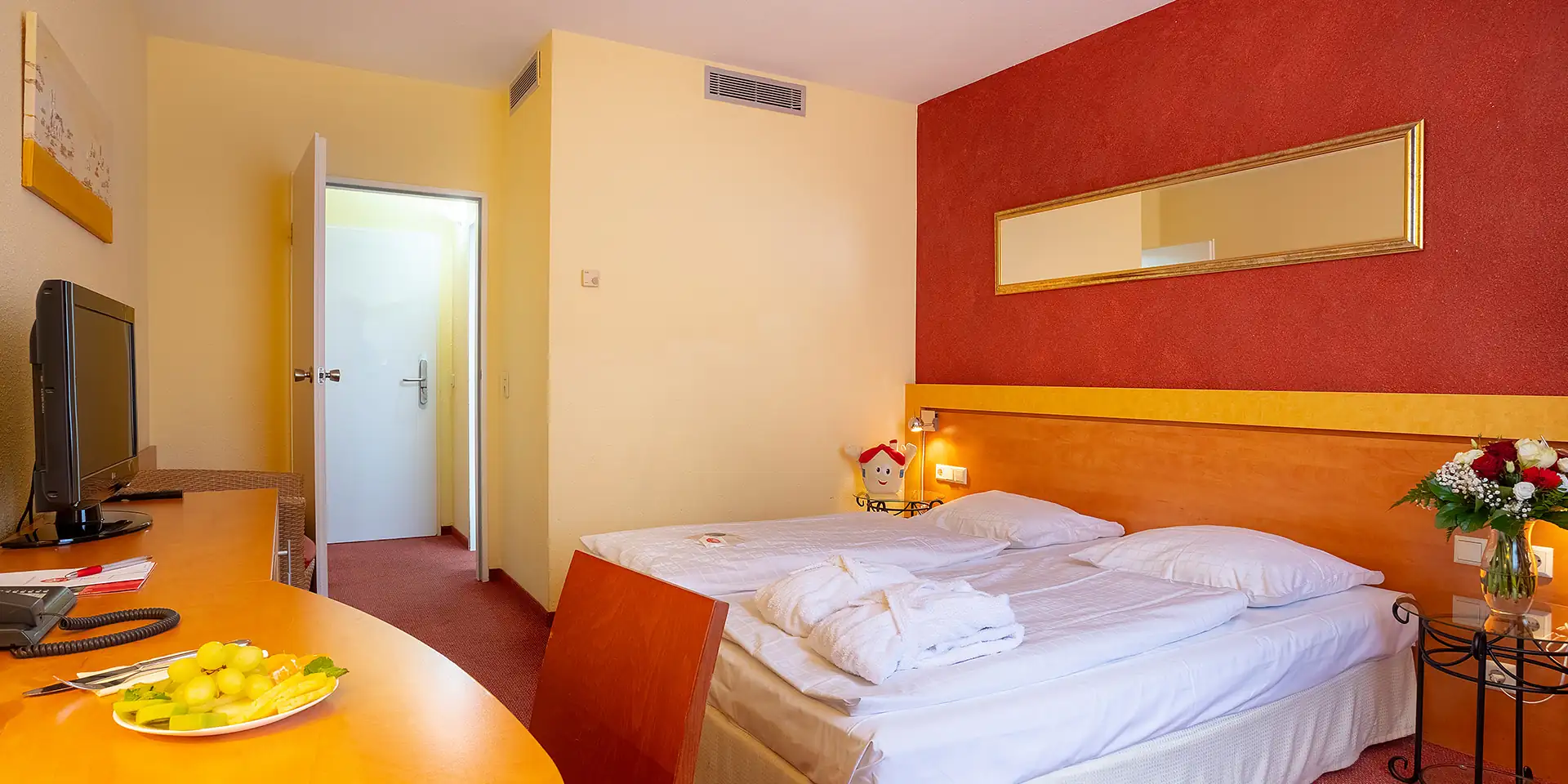 Hotelzimmer mit Doppelbett, Nachttischlampen, Fernseher auf Kommode, Stuhl, Blumenstrauß auf kleinem Tisch und offenem Türbereich