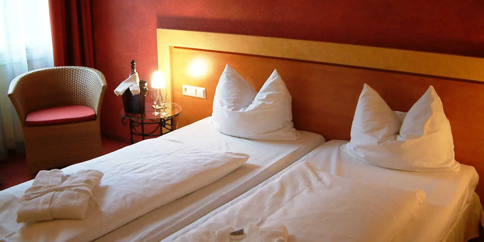 Hotelzimmer mit zwei Einzelbetten, Nachttisch mit Lampe und Sessel neben Fenster mit Vorhang