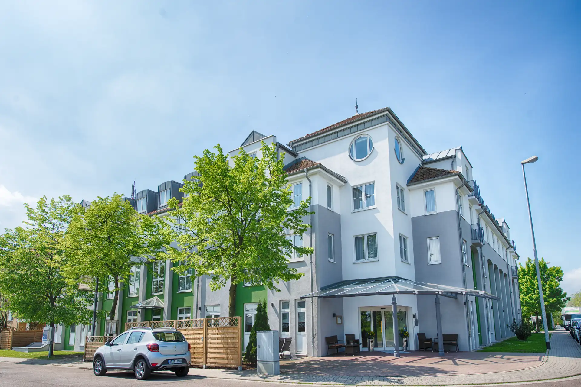 Achat Hotel Leipzig Messe, Außenansicht mit weißer, grauer und grüner Fassade im Sommer mit grünen Bäumen gesäumt.
