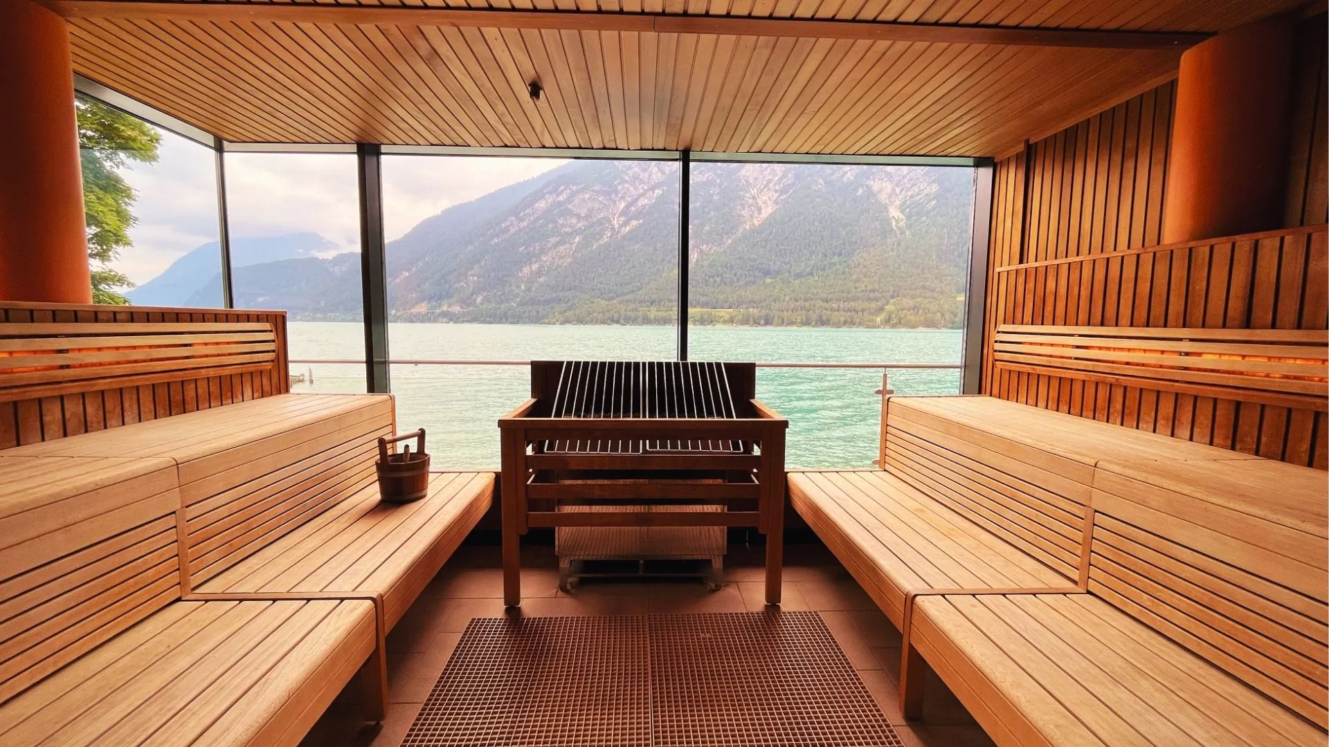 aja Fürstenhaus am Achensee Sauna aus hellem Holz mit großen Fenstern, durch die man Blick auf den Achensee und die Berge dahinter hat. Helles Tageslicht schein in die Sauna.