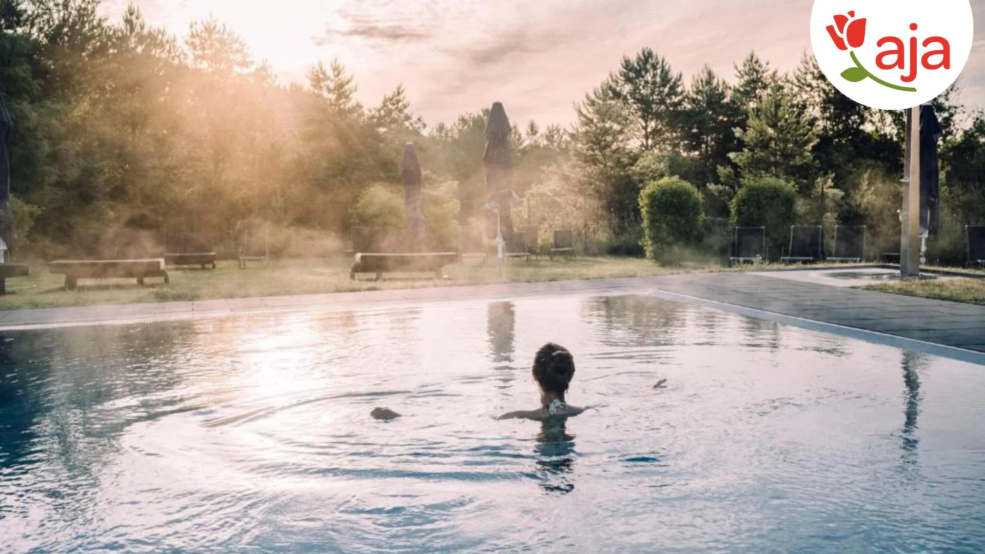 Ein idyllischer Außenpool im aja Resort Bad Saarow: Umgeben von grüner Natur und blauen Sonnenschirmen lädt der Pool mit glitzerndem Wasser zu einem entspannten Start in den Tag ein. Die sanften Sonnenstrahlen am Morgen sorgen für eine ruhige und erfrischende Atmosphäre. -mit Logo