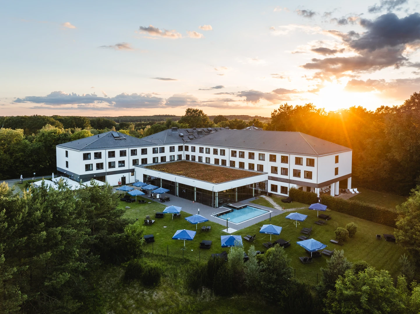 Das aja Resort Bad Saarow präsentiert sich inmitten idyllischer Natur, umgeben von grünen Wäldern und weitläufigen Wiesen. Der moderne Bau überzeugt mit einer großzügigen Gartenanlage, gemütlichen Liegebereichen und einem einladenden Außenpool. Der Sonnenuntergang taucht das Resort in eine warme, beruhigende Atmosphäre – perfekt für Erholung und Entspannung.