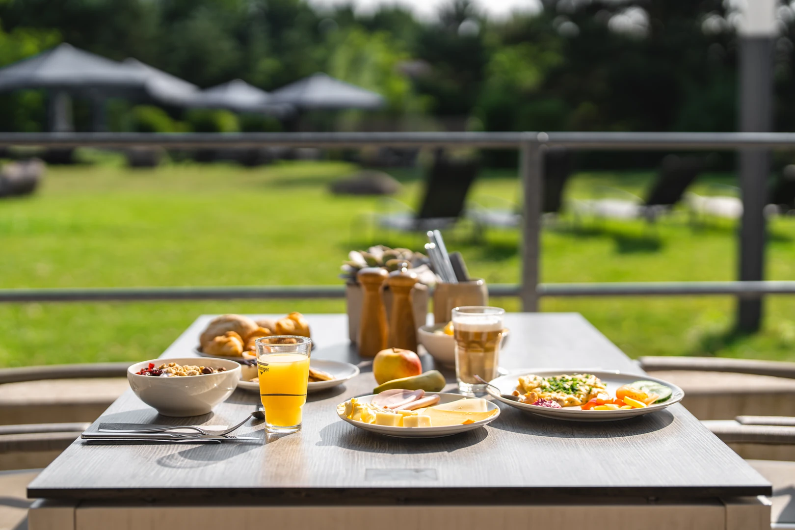 Ein Frühstück im Freien im aja Resort Bad Saarow: Auf einem stilvoll gedeckten Tisch werden frische Speisen wie Croissants, Obst, Käse und ein Glas Orangensaft serviert. Im Hintergrund erstreckt sich eine grüne Gartenlandschaft, die eine ruhige und entspannte Atmosphäre schafft.