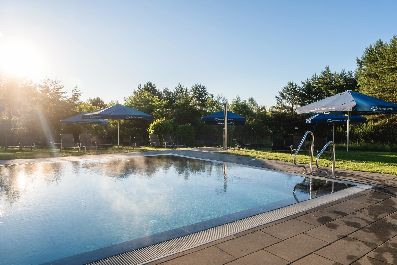 Ein idyllischer Außenpool im aja Resort Bad Saarow: Umgeben von grüner Natur und blauen Sonnenschirmen lädt der Pool mit glitzerndem Wasser zu einem entspannten Start in den Tag ein. Die sanften Sonnenstrahlen am Morgen sorgen für eine ruhige und erfrischende Atmosphäre.