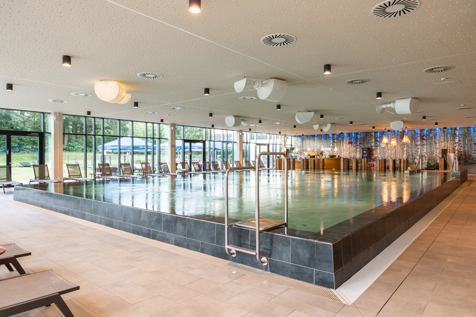 Das großzügige Hallenbad im aja Resort Bad Saarow bietet eine moderne, helle Atmosphäre mit bodentiefen Fenstern, die den Blick auf die grüne Natur freigeben. Bequeme Liegen laden zum Entspannen ein, während das stilvolle Design und die sanfte Beleuchtung eine einladende Umgebung schaffen.