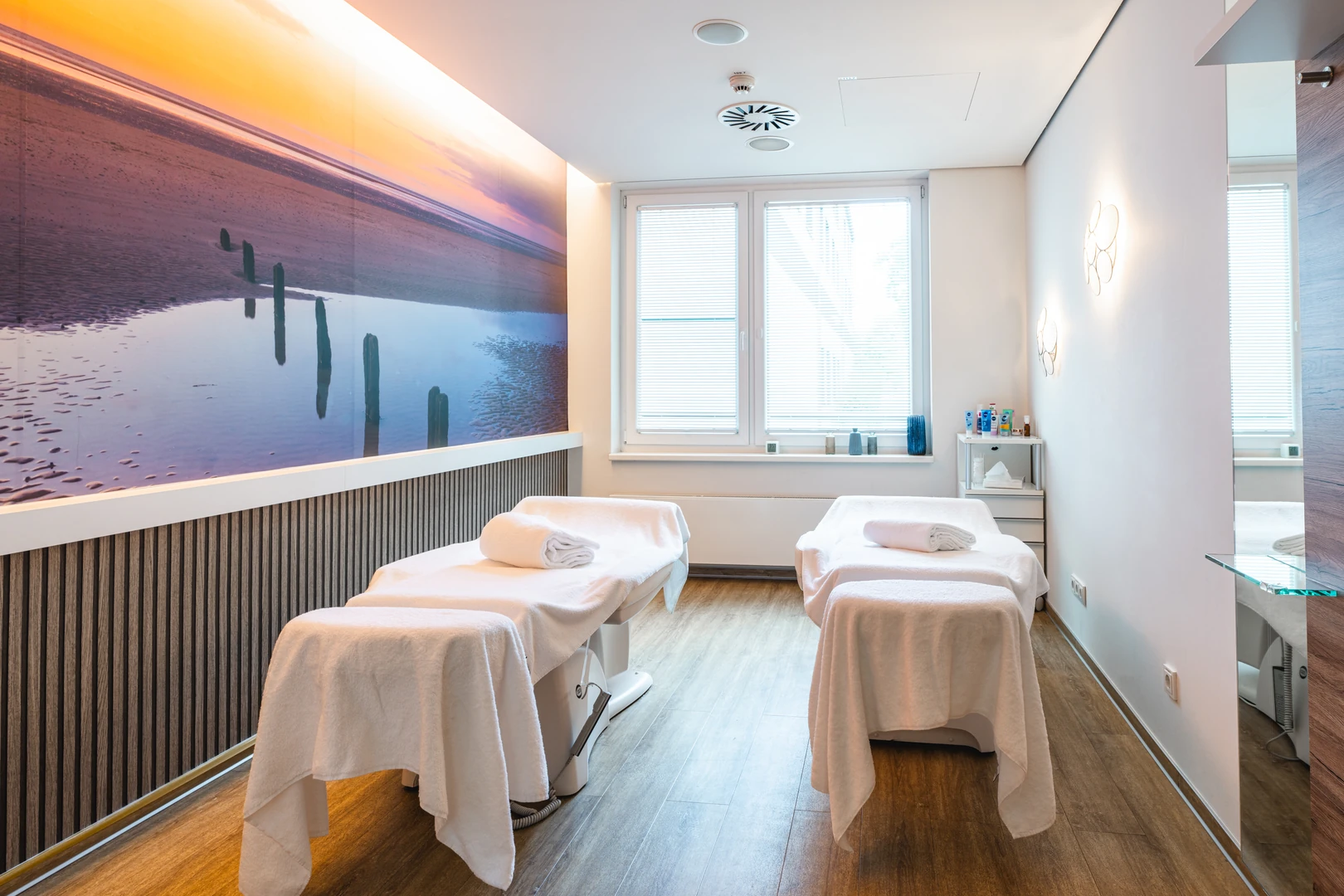 Der stilvolle Massageraum im aja Resort Bad Saarow lädt mit beruhigendem Ambiente und hochwertigem Design zur vollkommenen Entspannung ein. Die stimmungsvolle Wandgestaltung mit Naturmotiven und die modernen Behandlungsliegen schaffen eine perfekte Atmosphäre für wohltuende Wellness-Momente.