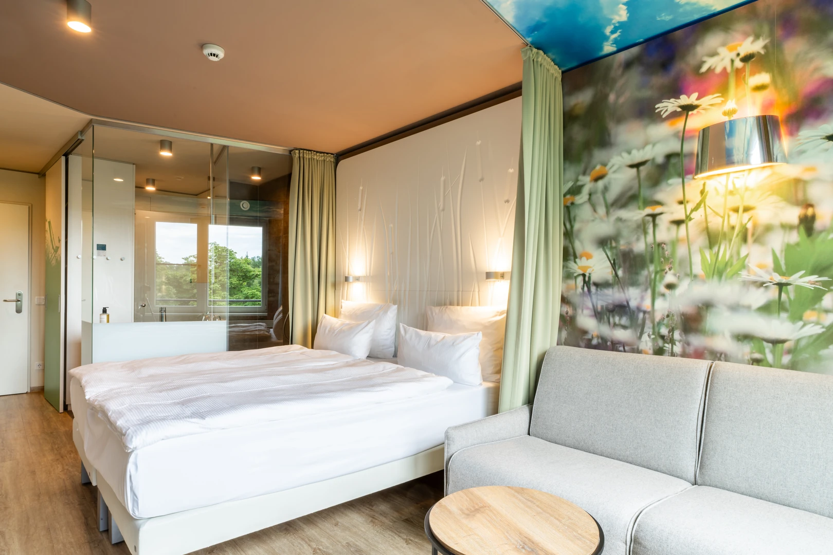 Das Zimmer im aja Resort Bad Saarow kombiniert modernes Design mit einer natürlichen Atmosphäre. Die gläserne Dusche und die harmonische Farbgestaltung schaffen einen Ort zum Wohlfühlen. Die liebevollen Details wie die floralen Wandmotive runden das stilvolle Ambiente ab.