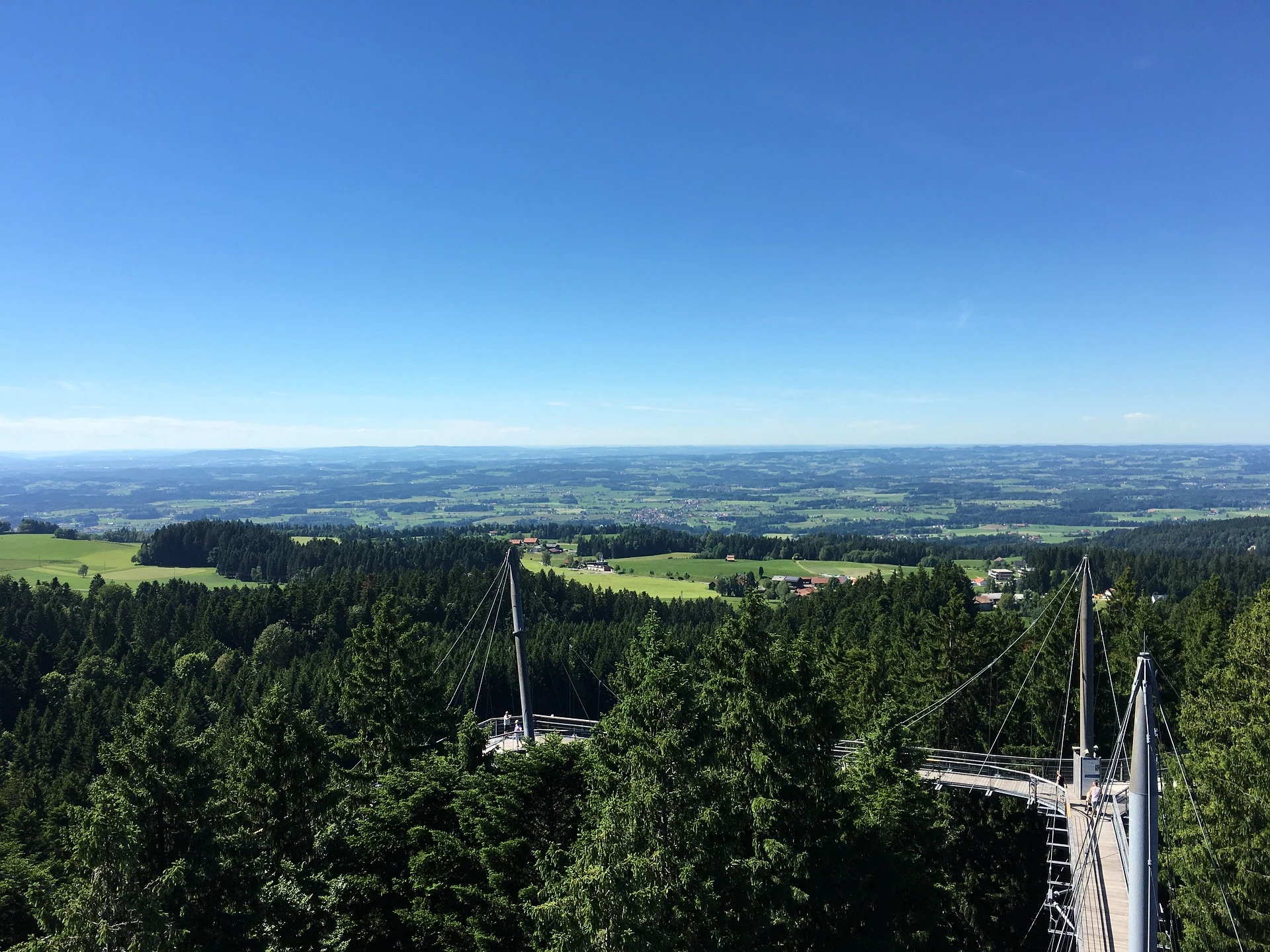 Das Bild zeigt den Baumkronenweg im Allgäu, der den Besuchern einen atemberaubenden Blick über die umliegenden Wälder und das weite Land bietet. Der Weg führt über eine hochgelegte Brücke, die zwischen den Bäumen schwebt und die Aussicht auf die unberührte Natur in der Region ermöglicht. Der klare Himmel und das grüne Panorama schaffen eine friedliche Atmosphäre und machen den Baumkronenweg zu einem unvergesslichen Erlebnis für Naturfreunde und Wanderer.