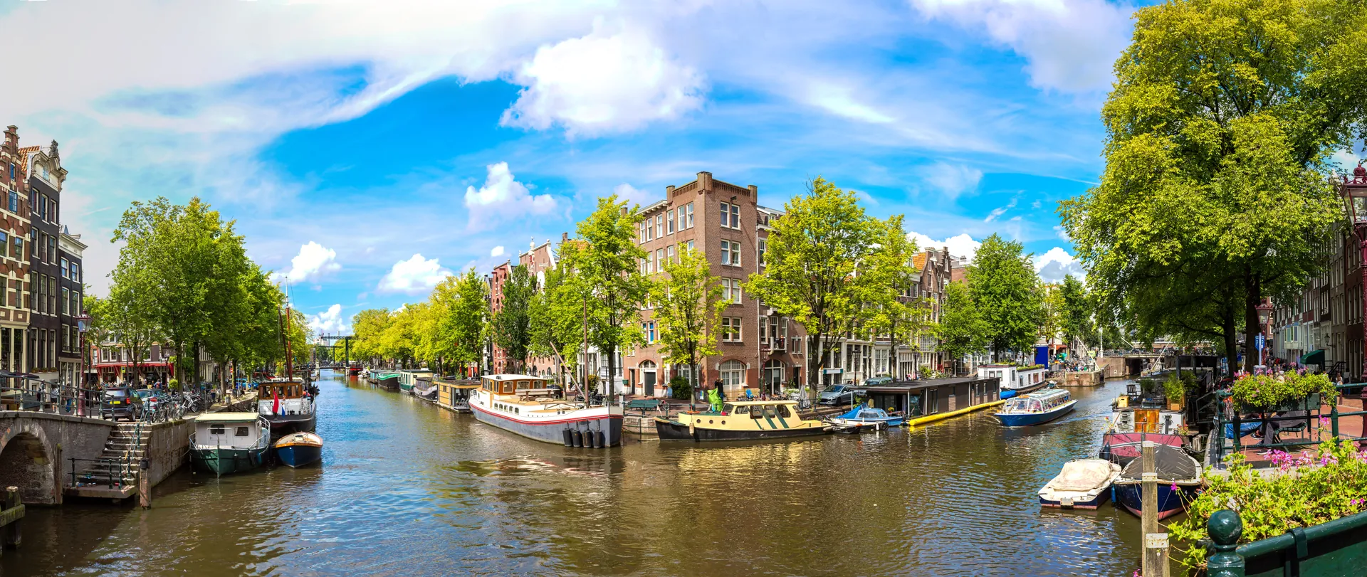Amsterdam Kanalbrücke bei Sonnenschein, umgeben von malerischen Grachten und zahlreichen angelegten Booten sowie kleinen Schiffen. Die Brücke bietet einen schönen Blick auf die lebendige Atmosphäre des Wassers und die charakteristischen Häuser, die die Ufer säumen. Das klare Wetter verstärkt die Farben und das einladende Ambiente der Stadt.